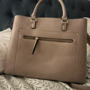 Beige handväska  - Snygg beige handväska med stilrena guldiga detaljer. Väskan har både handtag och axelrem, samt flera fack och dragkedjor för smart förvaring. Perfekt för dig som vill ha en klassisk och rymlig väska med stilren look.