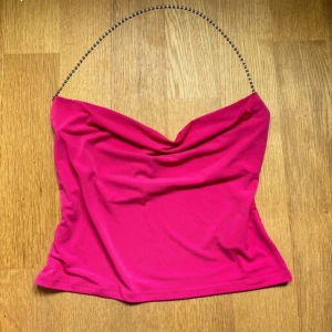 Rosa topp med strassband från NA-KD. M - Säljer en snygg rosa topp från NA-KD med glittrigt strassband som axelband. Toppen har draperad urringning och är figurnära med croppad passform. Perfekt för dig som vill sticka ut med en färgstark och trendig look. Storlek M