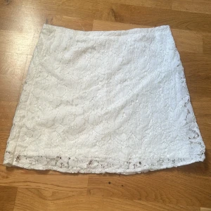 Vit spetskjol med kort längd - Säljer en vit kortkjol med vacker spetsdetalj. Kjolen har en rak och enkel form som ger en clean look, perfekt för dig som gillar stilrena plagg med lite extra touch. Spetsen gör den extra feminin och lätt att matcha med olika toppar.