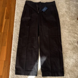 Svarta pleated chinos från Gant - Snygga svarta chinos från Gant i modellen Pleated Volume. Byxorna har vida ben, pressveck och klassisk passform med hällor för bälte. Tillverkade i bomull och perfekta för en clean och stilren look.