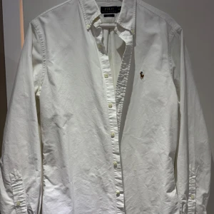 Vit skjorta Polo Ralph Lauren slim fit - Klassisk vit skjorta från Polo Ralph Lauren i slim fit-modell. Skjortan har knappar framtill, button down-krage och den ikoniska broderade loggan på bröstet. Tillverkad i bomull och har långa ärmar. Perfekt för en clean och stilren look.