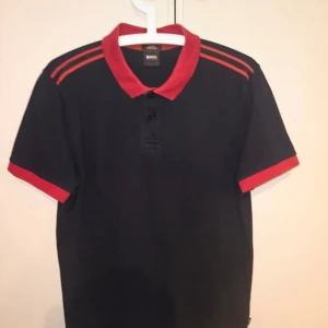 Svart och röd pikétröja från BOSS - Snygg pikétröja från BOSS i svart med röda detaljer på krage, axlar och ärmslut. Tröjan har klassisk pikékrage, tre knappar framtill och korta ärmar. Materialet är bomull och modellen är slim fit, perfekt för dig som gillar stilrena och sportiga vibes.