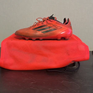 F50 ELITE - Säljer ett par röda Adidas F50 fotbollsskor med svarta detaljer och klassiska tre ränder på sidan. Skorna har snörning, platt sula med dobbar och är tillverkade i syntetmaterial för lätt känsla på planen. Perfekta för dig som vill sticka ut på fotbollsplanen. Får även med adidas påsen som tillhör skorna