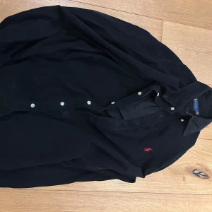 Svart manchesterskjorta Ralph Lauren - Snygg svart manchesterskjorta från Ralph Lauren med klassisk krage och knäppning framtill. Skjortan har det ikoniska röda broderade Polo-loggan på bröstet och är långärmad. Perfekt för dig som gillar stilrena och tidlösa plagg.