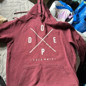 Vinröd hoodie från Dope, storlek S - Säljer en vinröd hoodie från Dope i storlek S. Tröjan har stor vit print på framsidan med DOPE-logga, snörning i huvan och en klassisk känguruficka. Perfekt för chill dagar och streetwear-stil. Materialet är mjukt och känns som bomull.