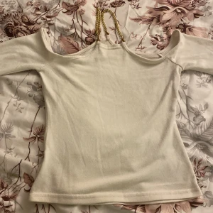 Offshoulder topp med guldkedjor - Snygg offshoulder topp i ljusbeige med unika axelband i guldfärgad kedja. Toppen har lång ärm och är figurnära i modellen. Perfekt för dig som vill sticka ut med coola detaljer och en clean look.