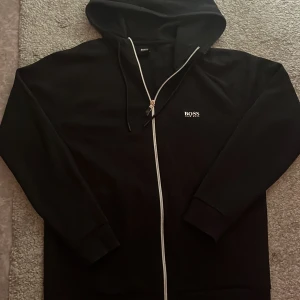 Svart hoodie från Hugo Boss - Svart hoodie från Hugo Boss med vit dragkedja och logga på bröstet och ryggen. Tröjan har huva med snörning och långa ärmar. Stilren design som passar perfekt till streetwear och chill dagar. Materialet känns mjukt och skönt mot huden.