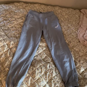 Ljusgrå/blå mjukisbyxor från Puma - Snygga ljusgrå/blåmjukisbyxor från Puma med resår i midjan och vid bensluten. Byxorna har en klassisk logga på låret och är tillverkade i mjukt material som är skönt att ha på sig. Perfekta för chill eller träning.