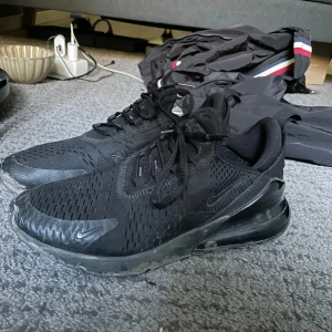 Svarta Nike Air Max 270 sneakers - Säljer ett par helsvarta Nike Air Max 270 sneakers i storlek 42. Skorna har en luftig mesh-överdel, synlig Air Max-dämpning i hälen och klassisk Nike-logga på sidan. Perfekta för dig som gillar sportig och stilren look.