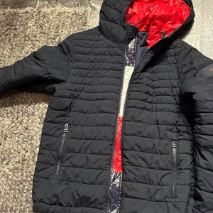 Svart pufferjacka från Tommy Hilfiger - Snygg svart pufferjacka från Tommy Hilfiger med röda och vita detaljer på insidan. Jackan har huva, två dragkedjefickor och quiltad design. 