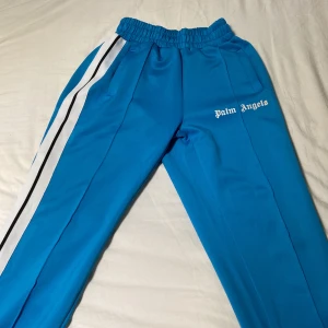 Palm Angels blåa trackpants - Blåa trackpants från Palm Angels med vita och svarta sidoränder, elastisk midja och dragkedjor vid bensluten. Byxorna har logga broderad på ena benet och är gjorda i ett mjukt syntetmaterial. Perfekta för en sportig och trendig look.