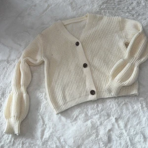 Beige stickad kofta med puffärm - Säljer en beige stickad kofta med v-ringning och bruna knappar framtill. Koftan har långa puffärmar med ribbade muddar och är croppad i modellen. 