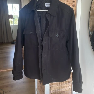 Svart overshirt från Weekday - Svart overshirt från Weekday med klassisk krage och två stora bröstfickor med knappar. Quiltad insida för extra komfort och värme. Långärmad modell med knappstängning framtill, perfekt för lager-på-lager-stil.