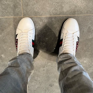 Gucci ace - Gucci Ace sneakers i vitt skinn med broderad röd och svart orm på sidan, klassiska gröna och röda ränder samt metallic grön och röd häl. Platt sula med unikt mönster undertill och snörning framtill. Perfekt för dig som vill sticka ut med lyxig streetstyle.