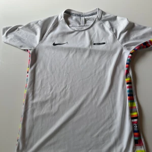 Vit Nike Mercurial Dri-Fit t-shirt - Snygg vit Nike Mercurial Dri-Fit t-shirt med korta ärmar och färgglada ränder längs sidorna. Tillverkad i ett lätt och ventilerande syntetmaterial som passar perfekt för fotboll eller träning. Diskret Nike-logga på bröstet och sportig design.