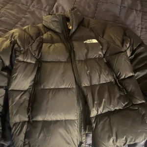 Svart dunjacka från The North Face - Säljer en svart dunjacka från The North Face, modell Hyalite Down. Jackan har en puffig design, hög krage och dragkedja framtill samt två sidofickor med dragkedja. Perfekt för kalla dagar och riktigt snygg streetstil. Helt oanvänd, lappen är fortfarande kvar. 