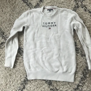 Vit stickad tröja Tommy Hilfiger - Vit stickad tröja från Tommy Hilfiger med broderad logga och text på bröstet. Klassisk rund halsringning och långa ärmar. Perfekt för dig som gillar stilrena och enkla plagg med en touch av premiumkänsla.