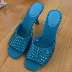 Blå glittriga klackar från H&M - Helt oanvända STORLEK 37. Snygga blå klackar från H&M med glittriga stenar över foten. Skorna har öppen tå, fyrkantig form och hög klack. Perfekta för att sticka ut och ge outfiten extra bling.