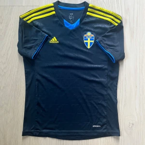 Svenska landslaget fotbollströja Adidas - Snygg marinblå fotbollströja från Adidas med Sveriges landslagsemblem på bröstet och gula Adidas-ränder på axlarna. Tröjan har korta ärmar med blå detaljer och är tillverkad i lätt, ventilerande polyester med Climacool-teknologi. Perfekt för dig som älskar svensk fotboll.