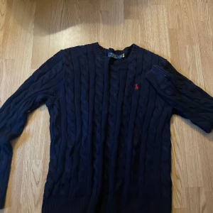 Marinblå stickad tröja Polo Ralph Lauren - Snygg marinblå stickad tröja från Polo Ralph Lauren med klassiskt kabelstickat mönster och röd broderad logga på bröstet. Rund hals och långa ärmar, perfekt för dig som gillar preppy stil. Passar dig som vill ha en tidlös och stilren look.