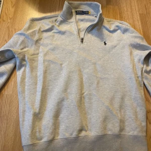 Ljusgrå half zip tröja Polo Ralph Lauren - Snygg ljusgrå långärmad tröja från Polo Ralph Lauren med klassisk broderad logga på bröstet. Tröjan har en stilren half zip-dragkedja och hög krage. Materialet är en mjuk blandning av bomull och återvunnen polyester, perfekt för chill dagar.
