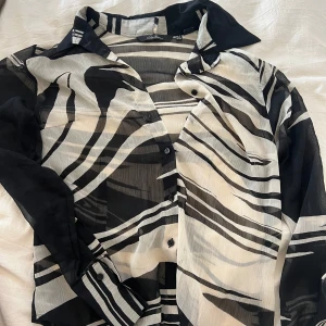 Svartvit zebramönstrad skjorta - Snygg transparent skjorta med svartvit zebramönster. Skjortan har klassisk krage, knappar framtill och långa ärmar med manschett. Perfekt för dig som gillar att sticka ut med coola mönster och lätt material.