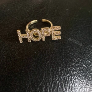 Guldfärgad ring med HOPE och stenar - Trendig guldfärgad ring med texten HOPE i stora bokstäver, täckta av glittrande stenar. Justerbar storlek och perfekt statement-piece för dig som vill sticka ut med ett positivt budskap.