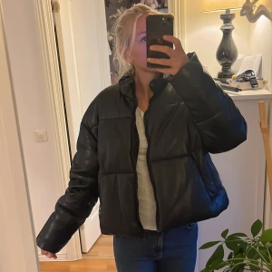 Svart pufferjacka i skinnimitation - Säljer en svart pufferjacka med oversized passform och dragkedja framtill. Jackan har en clean look med stora quiltade partier och är gjord i skinnimitation som ger en snygg glansig finish. Perfekt för dig som vill ha en trendig och varm jacka till hösten eller vintern.