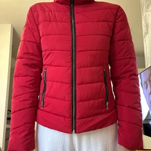 Röd pufferjacka med dragkedjor - Säljer en röd pufferjacka med svarta detaljer och två sneda fickor med dragkedjor. Jackan har quiltad design, hög krage och hel dragkedja framtill. Perfekt för kyliga dagar och ger en cool vibe till din outfit.   Jag bär storlek S och den passar