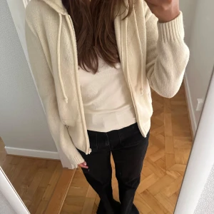 Beige stickad cardigan jacka med huva Brandy Melville - Mysig beige stickad cardigan jacka med huva och dragkedja framtill. Jackan har ribbade muddar, dragsko i huvan och två fickor. Bra kvalitet, 90% ull. Helt oanvänd, inga noppor.