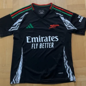 Helt ny oanvänd Arsenal svart kortärmad(priset kan diskuteras)Adidas tröja - Svart Arsenal fotbollströja från Adidas i slim fit med korta ärmar. Tröjan har gröna och röda ränder på axlarna, vitt tryck med Emirates Fly Better och Arsenal-logga i rött. Materialet är lätt polyester med vita grafiska detaljer på sidorna och Aeroready-teknologi.