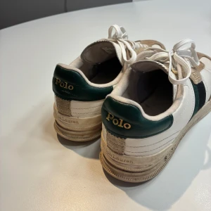 Polo Ralph Lauren sneakers i skinn och mocka - Snygga sneakers från Polo Ralph Lauren med vit skinnbas, beige mockadetaljer och gröna samt svarta inslag på sidan och hälen. Klassisk rund tå, platt sula och vita snören. Märkeslogga i guld på hälen och yttersulan. Perfekt för dig som gillar retrostil och streetwear.