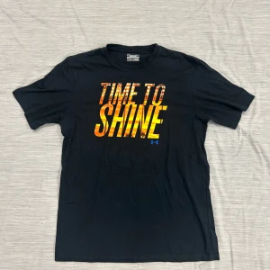 Svart t-shirt Under Armour Time To Shine - Svart t-shirt från Under Armour med trycket 'TIME TO SHINE' i orange och gul ton över bröstet. Klassisk rund hals och korta ärmar. Tillverkad i bomull med HeatGear-teknologi som håller dig sval. Perfekt för dig som gillar sportig och cool stil.