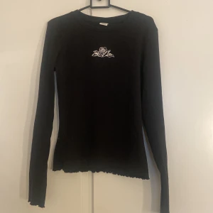 Svart långärmad tröja med ros från Lindex - Svart långärmad tröja från Lindex med vit ros på bröstet. Tröjan är ribbad. Perfekt för dig som gillar stilrena plagg med en liten detalj.
