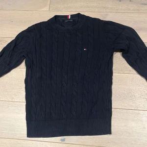 Svart kabelstickad tröja Tommy Hilfiger - Svart kabelstickad tröja från Tommy Hilfiger med rund hals och diskret logga på bröstet. Tröjan har ribbade muddar vid ärmslut och nederkant, samt långärmad passform. Perfekt för dig som gillar klassisk stil med en modern touch.