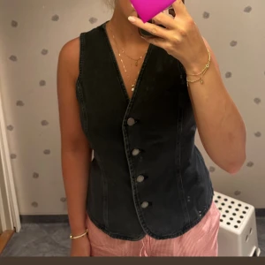 Svart jeansväst med knyt i midjan - Säljer en svart jeansväst från Mango med snygg v-ringning och knappar framtill. Västen har en figurnära passform och knytband i midjan för extra stilpoäng. Perfekt att styla över en t-shirt eller skjorta för en cool look💕