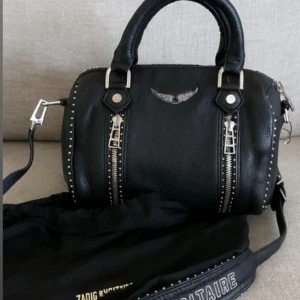 Svart handväska från Zadig & Voltaire - Cool svart handväska från Zadig & Voltaire med silverfärgade detaljer, dubbla dragkedjor framtill och dekorativa nitar längs kanterna. Väskan har både handtag och axelrem, samt ett snyggt emblem med vingar på framsidan. Dustbag och tagg medföljer. Vid snabb affär kan priset diskuteras! 