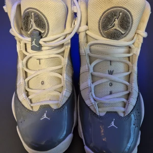 Nike Air Jordan 6 Rings vit/grå - Nike Air Jordan 6 Rings sneakers i vitt och grått med klassisk Jumpman-logga på plös och häl. Skorna har snörning, bred rem med AIR JORDAN-tryck och en blandning av skinn och textilmaterial. Snygg, sportig siluett med rund tå och platt sula.