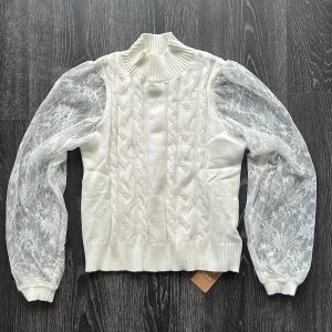 Vit stickad tröja med spetsärmar - Supersöt vit stickad tröja med höghalsad ribbad krage och långa transparenta spetsärmar med blommönster. Kroppen är kabelstickad och ärmarna har breda muddar. Perfekt för dig som vill ha något unikt och trendigt. Står L men det är helt fel mer som en S