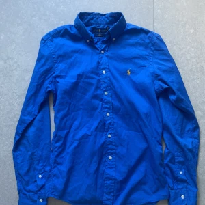 Blå slim fit skjorta från Ralph Lauren - Snygg blå skjorta från Ralph Lauren i slim fit-modell. Tillverkad i lätt bomullstwill med klassisk krage och knappar framtill. Ikoniska Polo-loggan broderad på bröstet i gult. Perfekt för dig som gillar stilrena och tidlösa plagg. Otroligt bra skick och pris går alltid att pruta!