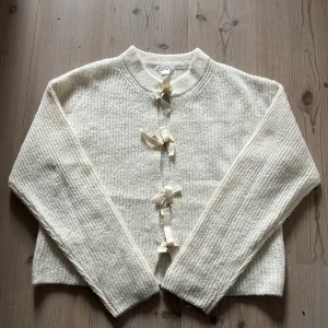 Krämvit kofta med rosettknäppning - Superfin krämvit kofta i stickad ullmix med söta rosetter frammtill. 