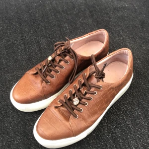Dasia skor brun - Snygga bruna sneakers i crocopräglat skinn med vita platta sulor och bruna snören. Guldiga öljetter och en liten metallcharm på snörningen ger extra detalj. Perfekta för dig som vill ha en stilren men ändå unik look. Anväd cirka 10 gånger. Säljer dessa för köpt samma skor fast i färgen svart❤️