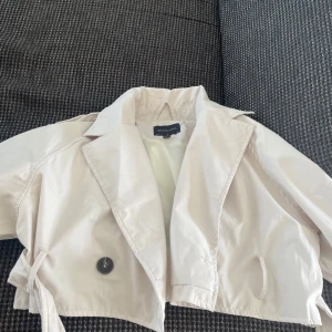 Beige croppad trenchcoat booho  - Snygg croppad trenchcoat från Booho i beige med klassisk krage, knappar och bälte i midjan. Jackan har långa ärmar med spänne vid mudden. Den har inga skador och aldrig använd. Är i storlek Xs men är lite oversized så skulle även passa de med lite större storlek beroende på vilken passform man vill