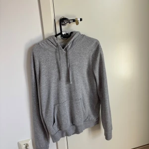 Grå hoodie med huva  - En basic grå hoodie med huva och snörning, samt en stor magficka framtill. Tröjan har långa ärmar och ribbade muddar vid ärmslut och nederkant. Perfekt för chill dagar eller när du vill ha något bekvämt och stilrent.