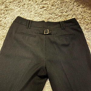 Svarta randiga kostymbyxor KappAhl - Snygga svarta kostymbyxor från KappAhl i storlek 42. Skulle snarare säga en storlek 40! Har inga defekter förutom en extra knapp som sitter löst, men den behövs inte då det finns två riktiga knappar. Coolt spänne bak!