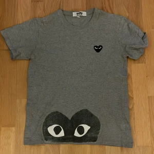 Grå Comme des Garçons Play t-shirt - Grå t-shirt från Comme des Garçons Play med klassiskt svart hjärta med ögon på bröstet och stor svart ögon-grafik längst ner. T-shirten är kortärmad och har en avslappnad passform. Perfekt för dig som gillar streetstyle och ikoniska prints.