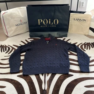 Mörkblå stickad tröja Polo Ralph Lauren - Snygg mörkblå stickad tröja från Polo Ralph Lauren med kabelstickat mönster och dragkedja framtill. Kragen har coola röda och svarta ränder på insidan. Perfekt för dig som gillar klassisk stil med en twist. Långärmad och med ribbade muddar för extra komfort.