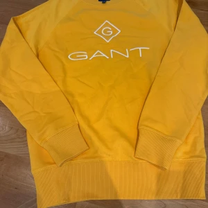Gul sweatshirt från GANT i bomull - Säljer en snygg gul sweatshirt från GANT i storlek S. Tröjan har ett stort vitt GANT-tryck på bröstet och ribbade muddar vid ärmslut och nederkant. Perfekt för dig som gillar färg och vill sticka ut lite extra. Tillverkad i mjuk bomull.