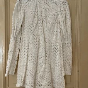 Vit kort klänning med spetsdetaljer - Supersöt vit klänning med broderade spetsdetaljer och långärmade puffärmar. Klänningen har en kort längd och är tillverkad i bomull, vilket gör den både luftig och bekväm. Perfekt för sommarens alla tillfällen och ger en romantisk vibe.
