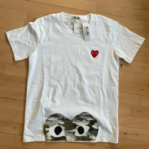 Vit Comme des Garçons T-Shirt  - Snygg vit t-shirt från Comme des Garçons Play med klassiskt rött hjärta med ögon broderat på bröstet och stort camo-tryck längst ner. Väldigt svår att få tag i! | Skick: 10 / 10, helt ny! | Marknadspris (säljs inte officiellt längre) ~1600 | Vårt pris: 799 